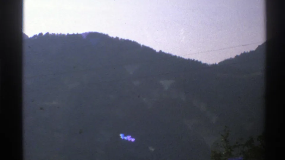 1976 Norway 8mm frame with a blue cast, hillside silhouette, faint lake surface, small trees at the bottom edge, and visible film grain.