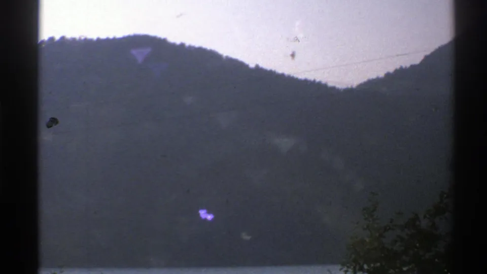 1976 Norway 8mm view of a forested mountain ridge seen through dark vertical window framing, with wires and a small bird silhouette.