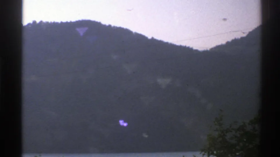 1976 Norway 8mm frame with a dark window edge, wooded mountain ridge, faint waterfront below, overhead wires, and purple artifacts.
