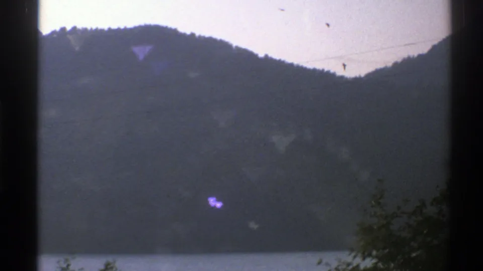 1976 Norway 8mm view of a wooded mountain above a faint waterfront, crossed by wires and framed by a dark window bar.