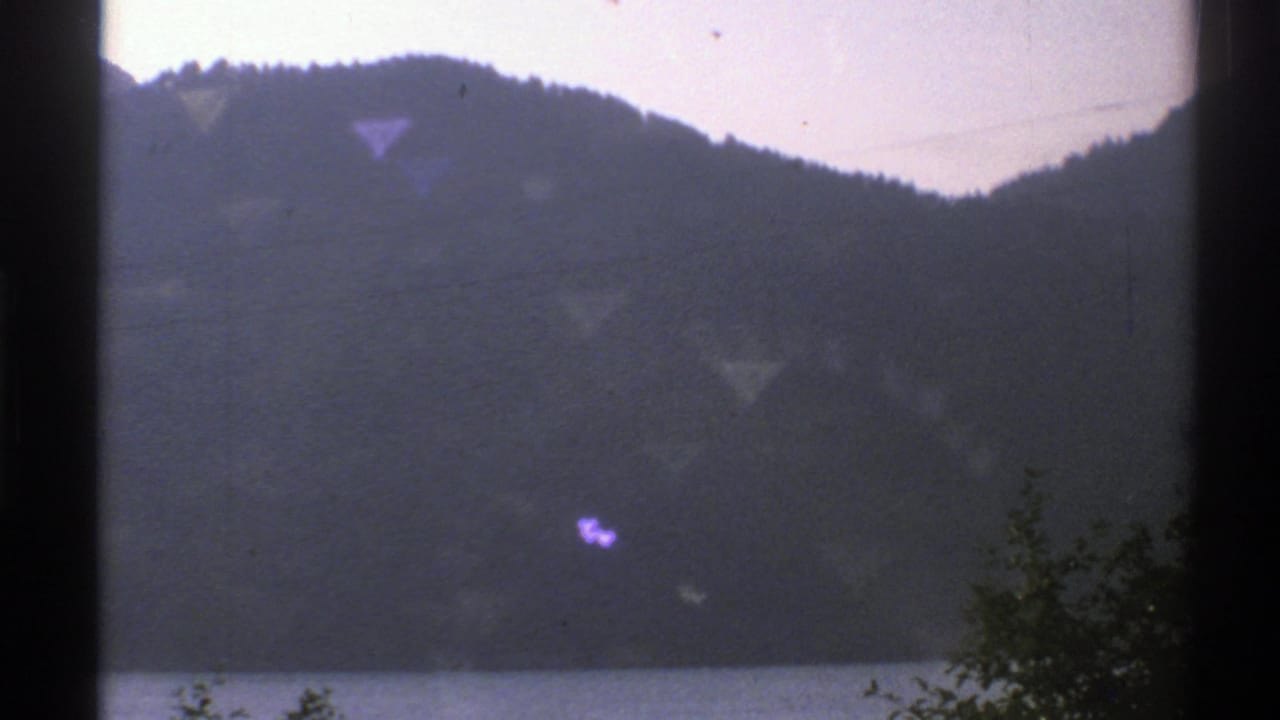 1976 Norway 8mm frame showing a wooded mountain beside calm water, with dark window-like borders and faint flare.