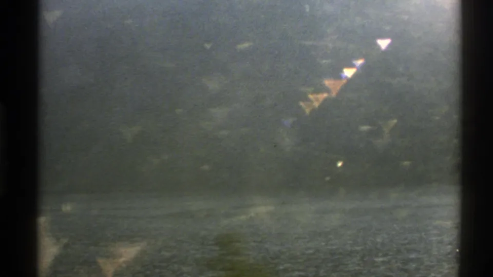 1976 Norway travel footage frame with rippled water, wooded shore, hazy hillside, and bright triangular flare artifacts.