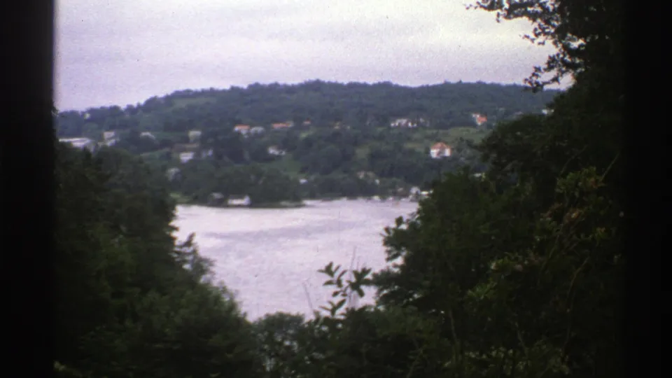 Norway 1976 8mm view over a lake toward wooded hills and scattered houses, framed by dark trees.