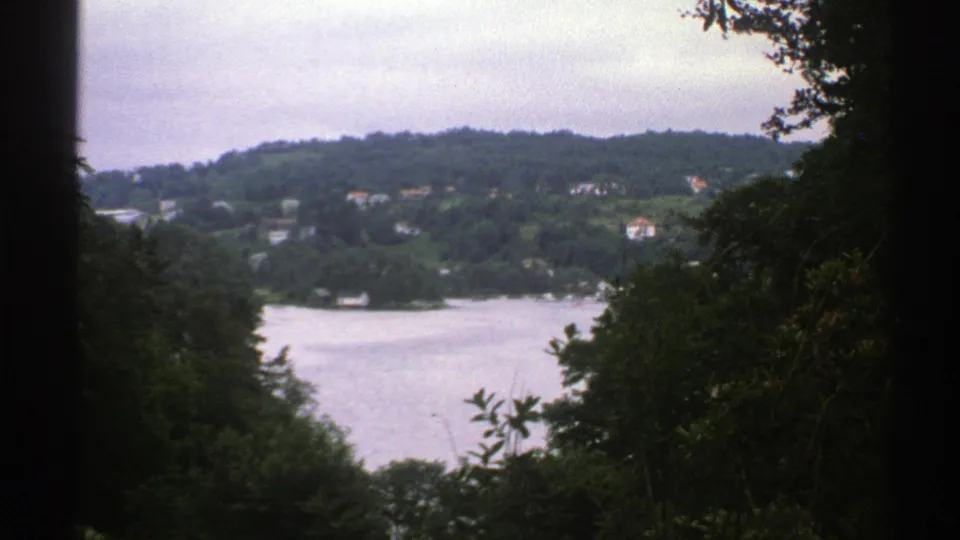 Norway 1976 home-movie frame showing a lake below wooded hills, with small distant houses and leafy branches in front.