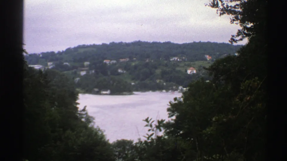 Norway 1976 8mm frame with a lake and wooded hillside settlement seen between dark foreground trees.