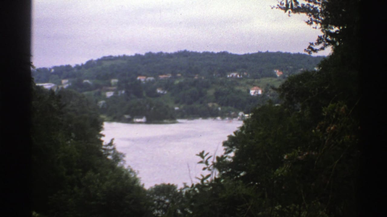 1976 Norway 8mm frame of a calm lake and wooded hillside with distant houses, viewed through shadowed trees.