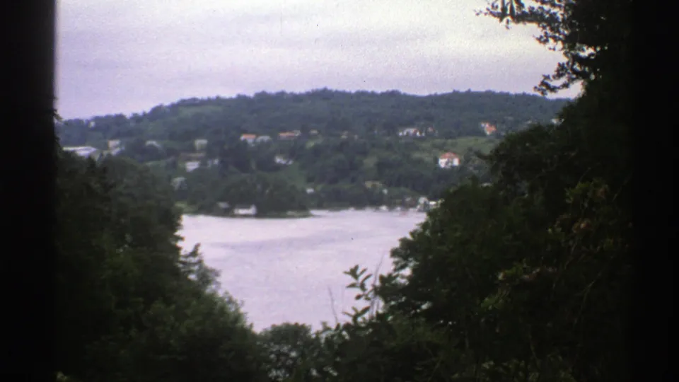1976 Norway 8mm frame of a calm lake and wooded hillside with distant houses, viewed through shadowed trees.
