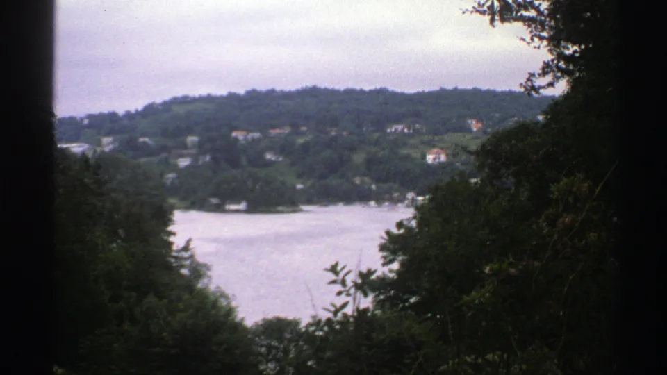 1976 Norway landscape frame showing a lake below wooded hills and small distant houses, bordered by dark foliage.
