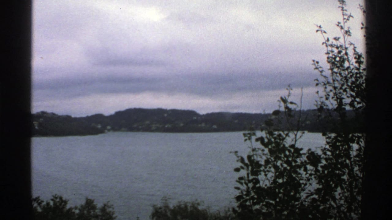 1976 Norway 8mm frame showing a blue-gray lake, low hills, scattered shoreline houses, dark foreground foliage, and heavy clouds.