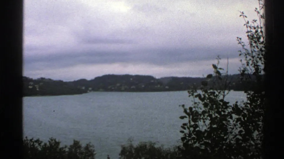 1976 Norway 8mm frame with muted lake water, wooded hills, small far-shore buildings, and dark foreground leaves under overcast light.