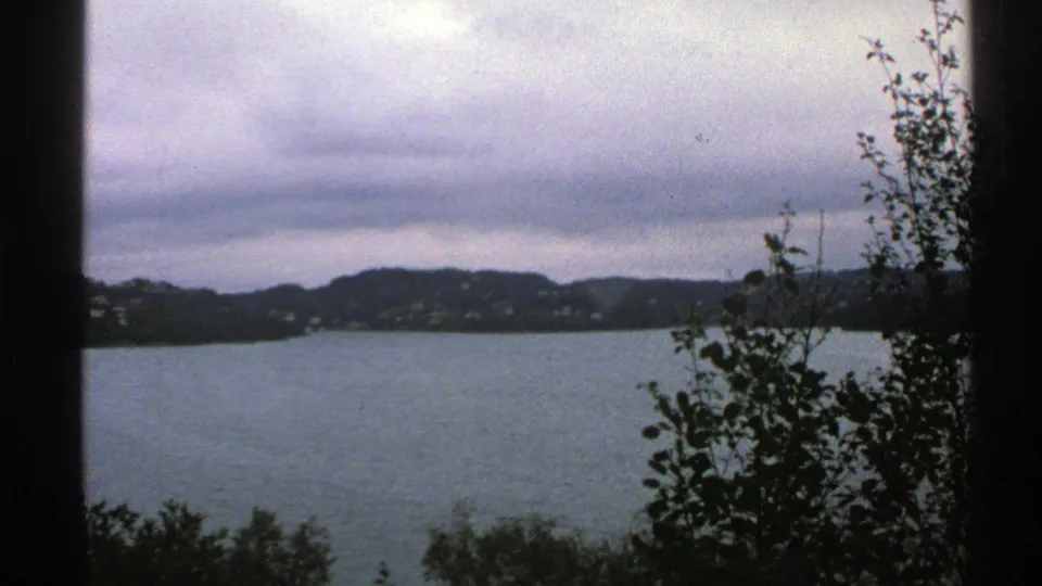 1976 Norway home-movie frame showing a blue-gray lake, low dark hills, scattered distant houses, and foreground trees silhouetted against the water.