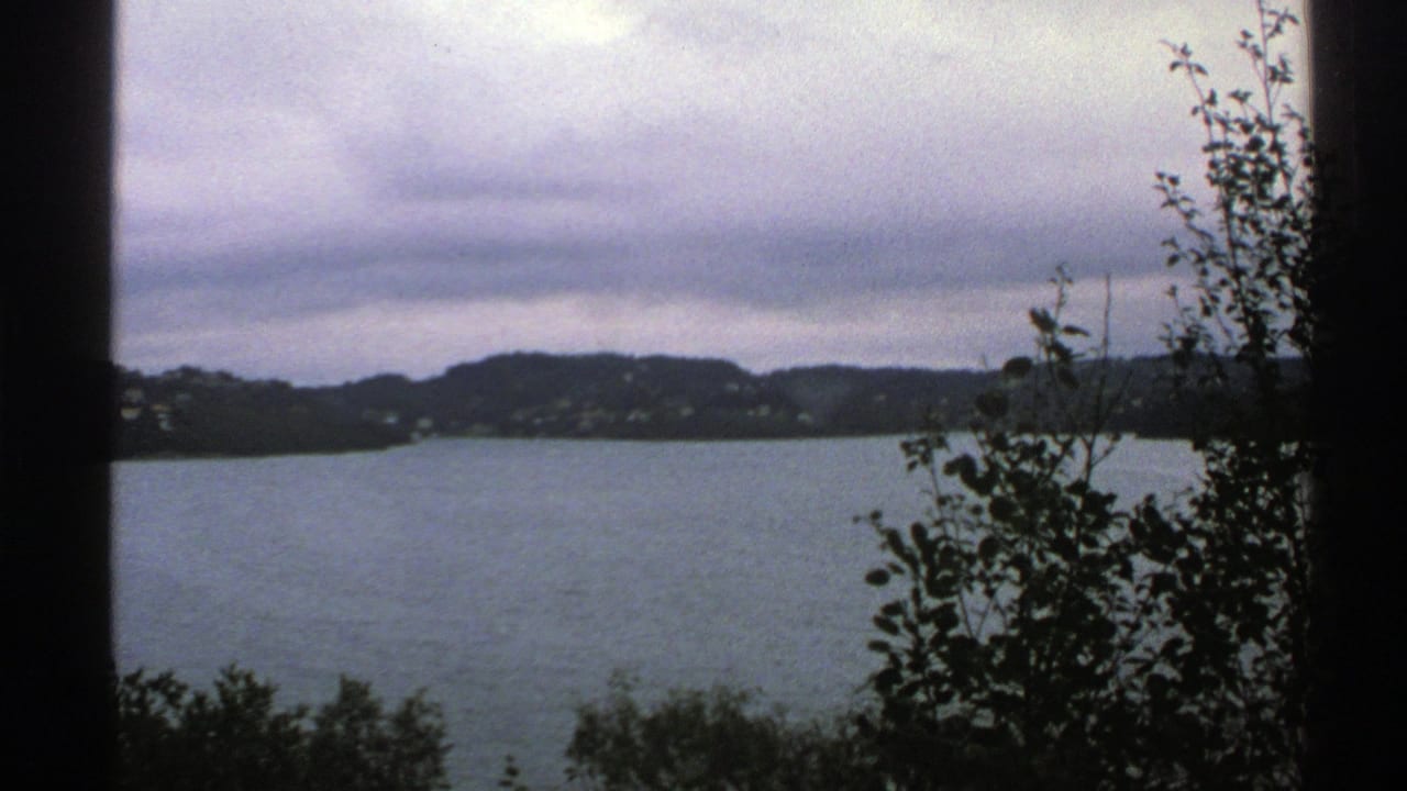 1976 Norway 8mm frame showing a calm lake, distant wooded hills, scattered houses, and foreground foliage viewed through a dark window frame.