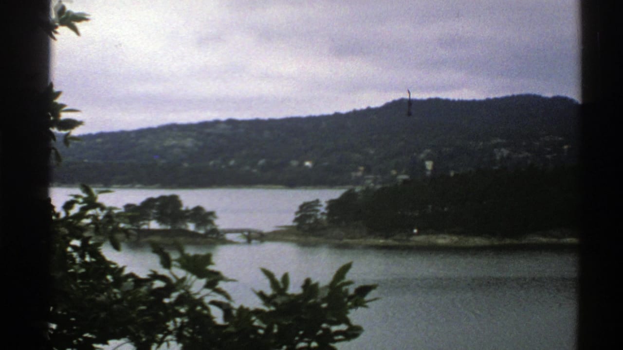 1976 Norway 8mm frame with a muted lake view, wooded hills, small distant houses, and dark window or post framing along the side.