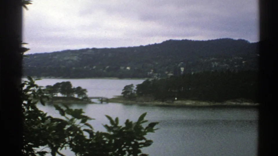 1976 Norway 8mm frame showing a lake inlet, tree-covered shoreline, distant hillside houses, and leafy foreground vegetation.