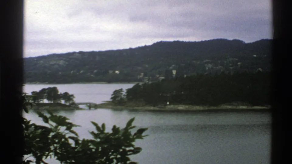 1976 Norway 8mm frame with calm water, a wooded point of land, distant hills, and foreground leaves beside dark window framing.