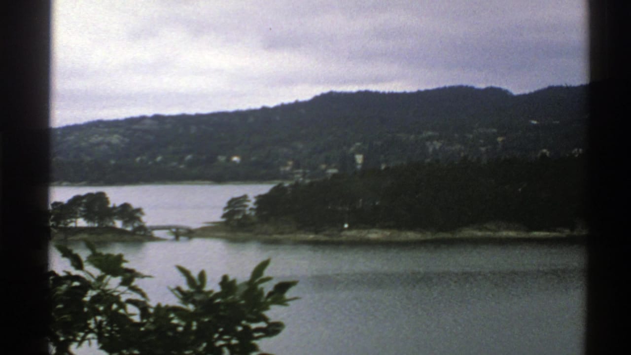 1976 Norway 8mm travel frame showing a fjord or lake, wooded shoreline, hills, and foreground leaves.
