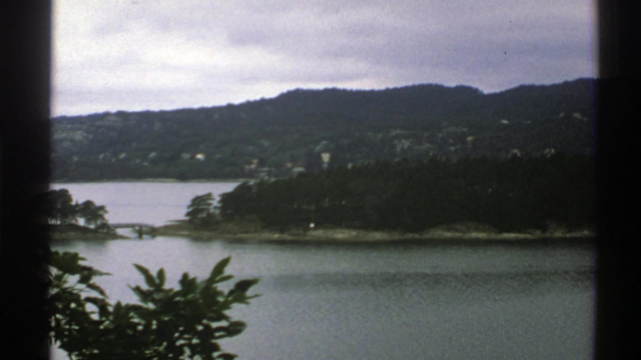 Elevated 1976 Norway landscape view across water to wooded hills and scattered distant buildings.