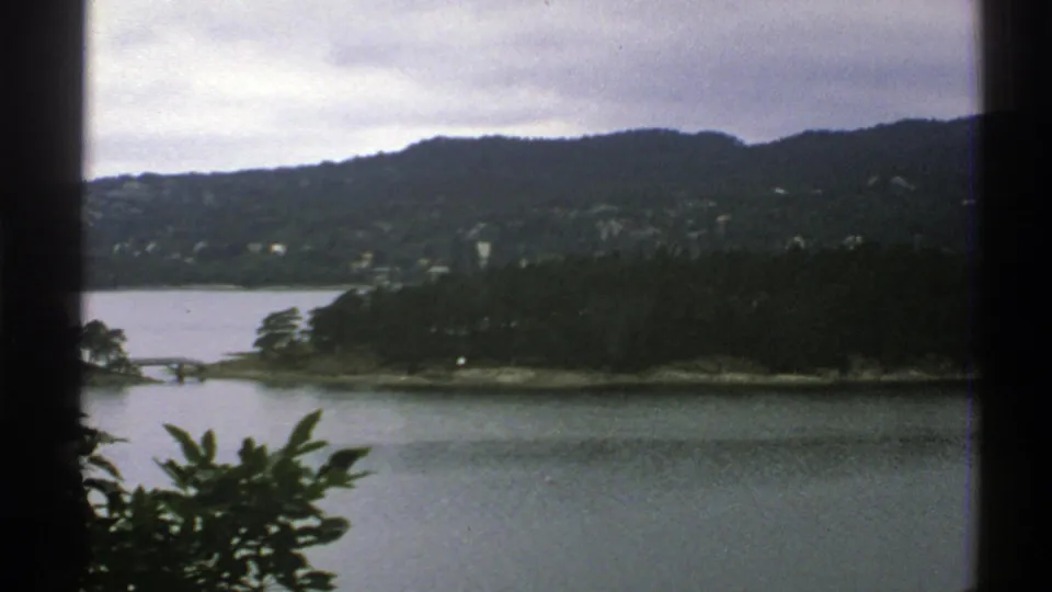 1976 Norway home-movie frame with water, wooded shore, hills, and soft 8mm color during the pan.
