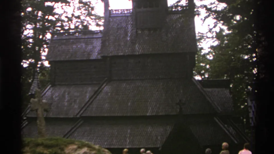 1976 Norway travel frame showing a dark wooden stave church roof among trees with visitors nearby.