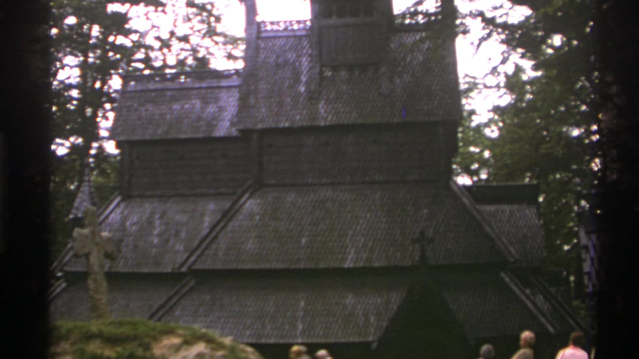 1976 Norway 8mm frame showing a dark timber stave church in a forested setting with a stone cross and visitors partly visible below.