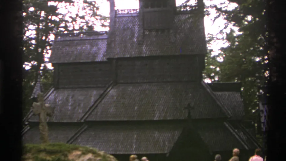 1976 Norway 8mm frame showing a dark timber stave church in a forested setting with a stone cross and visitors partly visible below.