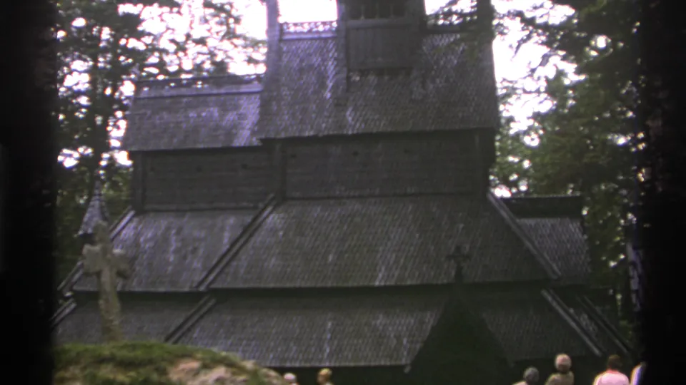 1976 Norway 8mm frame of a wooden stave church seen through trees, with distant visitors gathered near the entrance area.