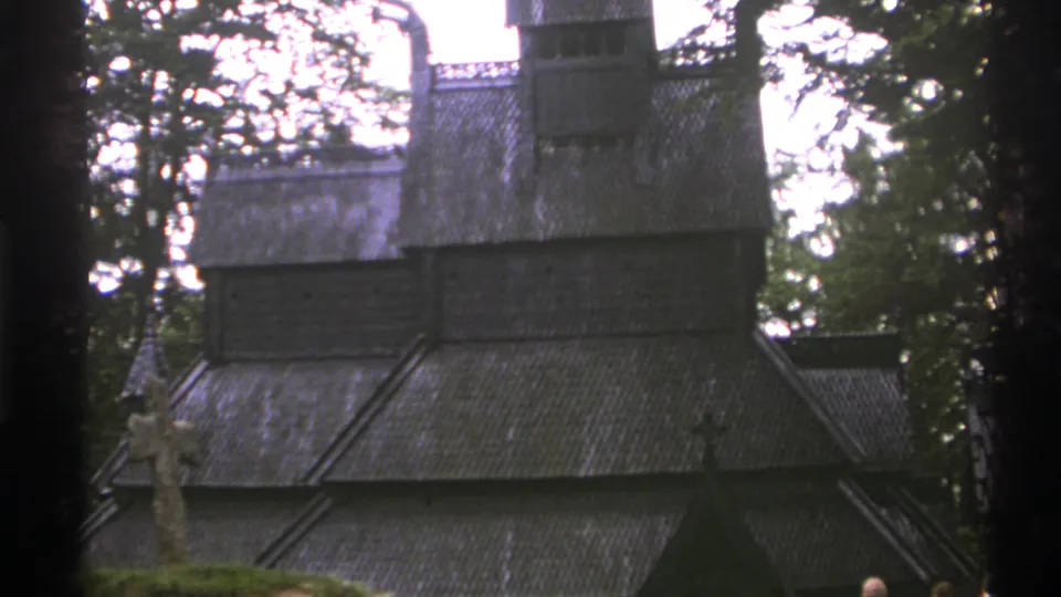 Soft 8mm view of a Norwegian stave church roof, tower, and carved-looking cross, with visitors partly visible at the lower frame edge.