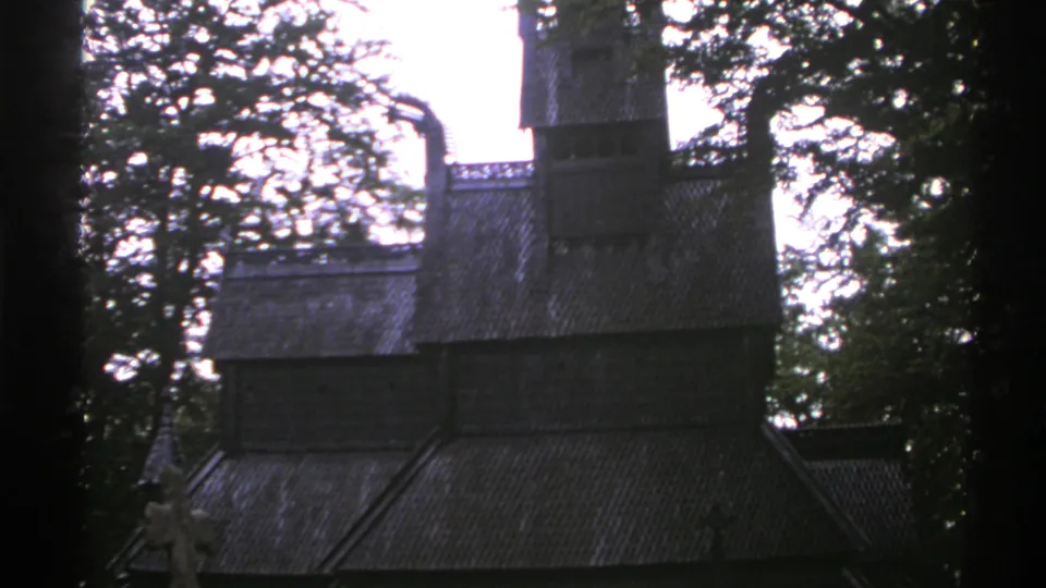 1976 Norway home-movie frame showing the stave church in darker exposure, with roof tiers, surrounding trees, and a cross at left.