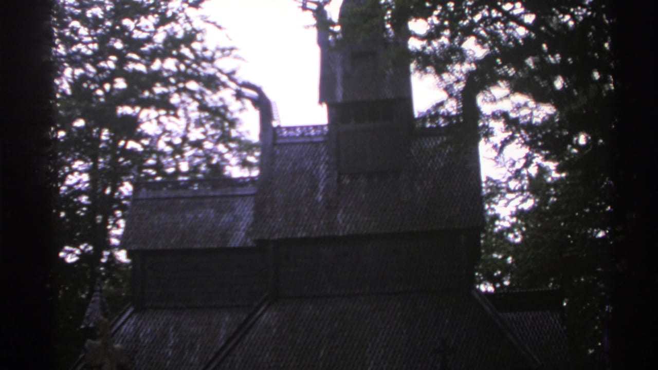 1976 Norway 8mm frame showing a dark wooden church-like structure partly obscured by trees against a pale sky.