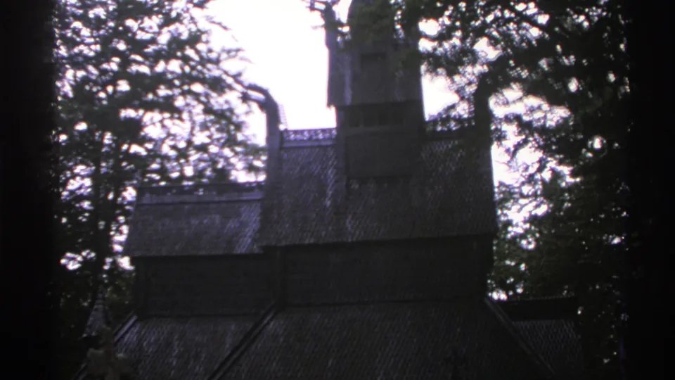 1976 Norway 8mm frame showing a dark wooden church-like structure partly obscured by trees against a pale sky.