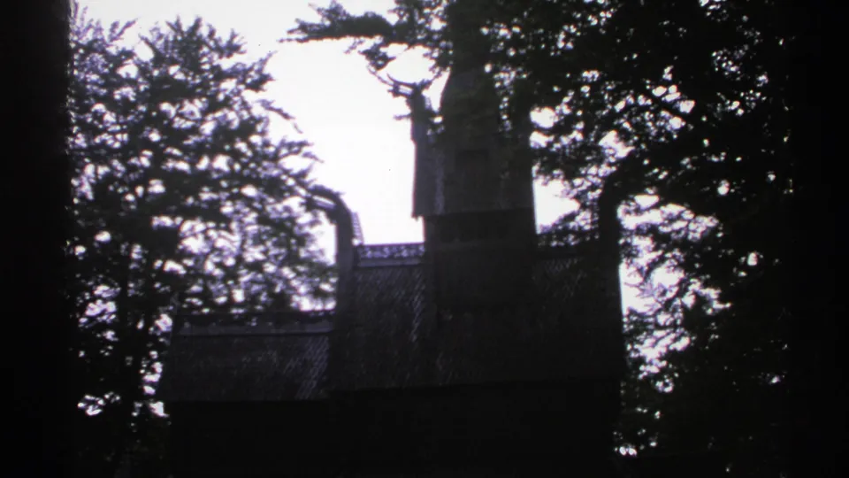 1976 Norway 8mm frame showing the upper part of a wooden church-like building behind dense trees.