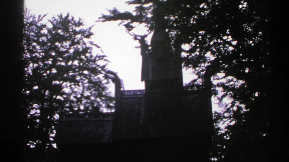 1976 Norway 8mm frame of a shadowed wooden landmark with a tower, seen through forest trees.