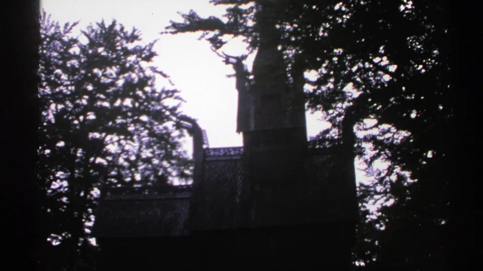Dark 8mm frame of a building partly hidden by trees, likely from the Norway 1976 travel reel.