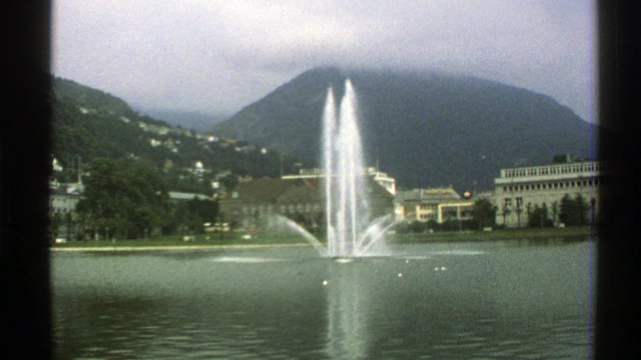 Lakeside fountain in front of buildings and a cloud-covered mountain.