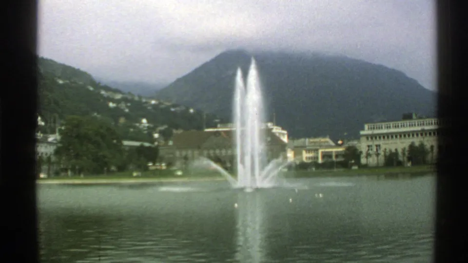 Vintage 8mm Norway lakeside view with a tall fountain, shoreline buildings, wooded slopes, and cloud along the mountain ridge.