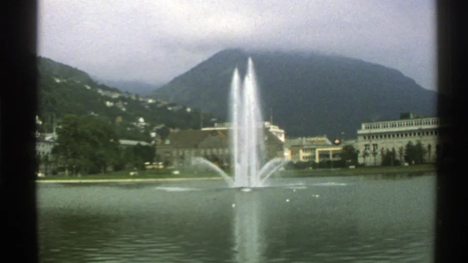 Norway 1976 home-movie frame of a lakeside fountain with calm water, civic-looking buildings, hillside trees, and low mountain cloud.