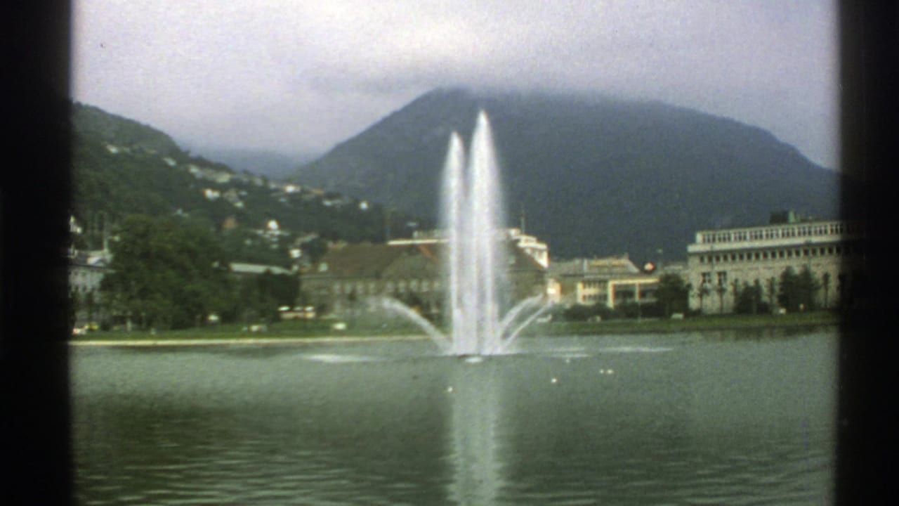 Lake fountain with mountains, shoreline buildings, trees, and cloudy sky.