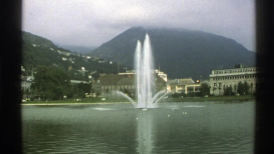 1976 Norway 8mm frame showing a fountain in a lake, misty mountains, trees, and waterfront buildings.