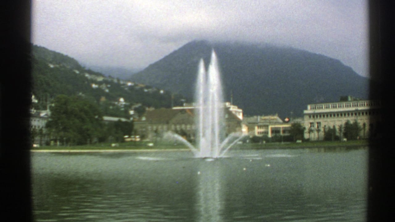 Lake fountain before misty mountains and a waterfront hotel building.