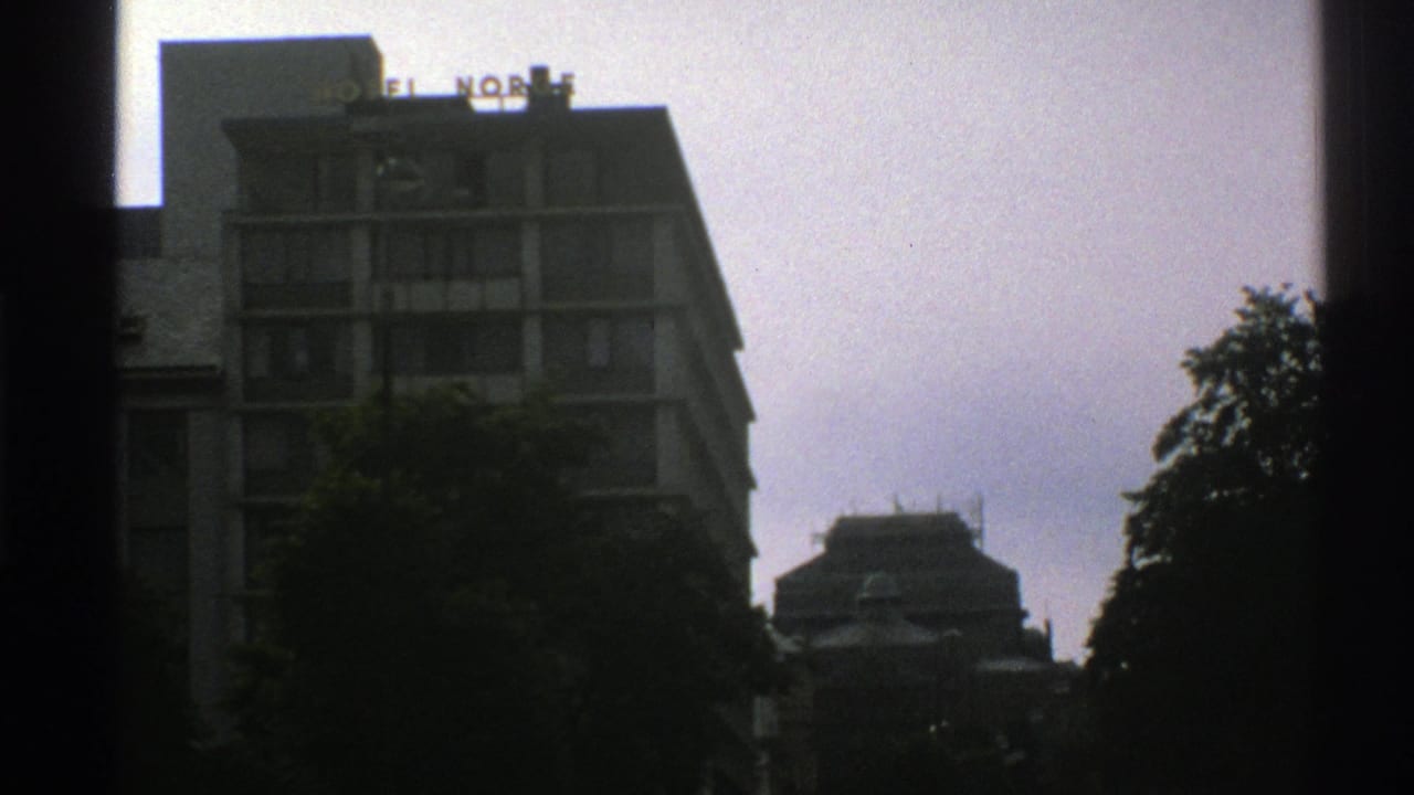 1976 Norway 8mm frame showing the Hotel Norge roof sign above a pale multi-story building, partly hidden by trees and window edges.