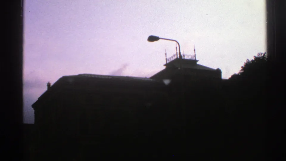 1976 Norway 8mm frame showing a dark building silhouette, streetlamp, rooftop details, trees, and overcast sky.