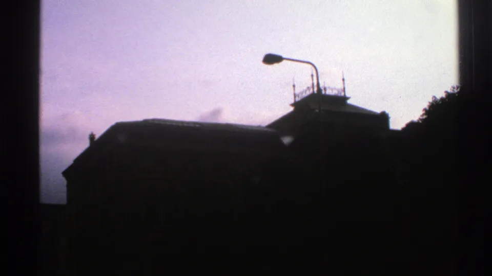 1976 Norway city-view frame with a streetlamp, silhouetted building roof, rooftop fixtures, trees, and pale overcast sky.