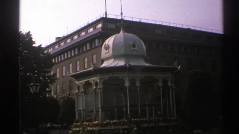 1976 Norway 8mm frame revealing a white domed gazebo, flowerbed, trees, and a larger historic-looking building behind it.