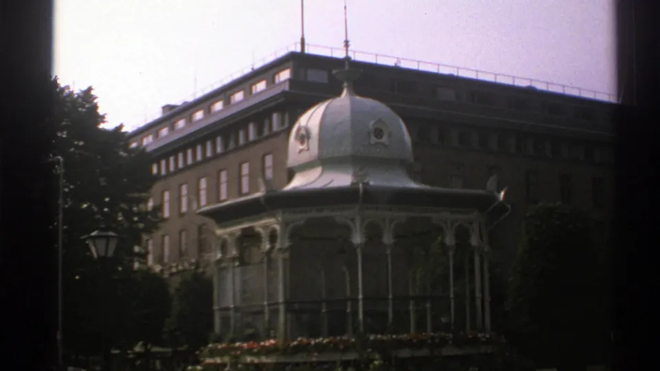 1976 Norway 8mm frame showing the white domed gazebo, surrounding flowers, trees, and the adjacent urban building facade.