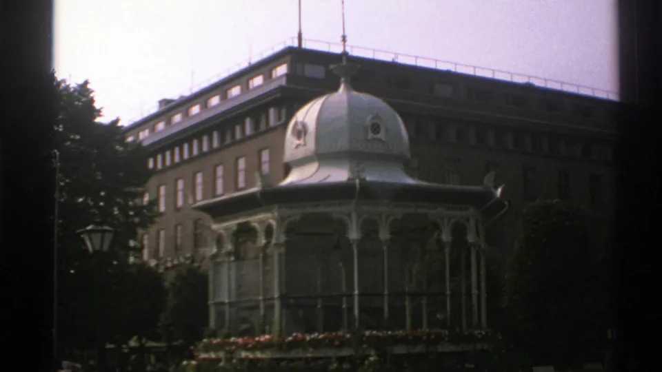 1976 Norway home-movie frame with the pavilion centered against a multi-story brick building and surrounding greenery.