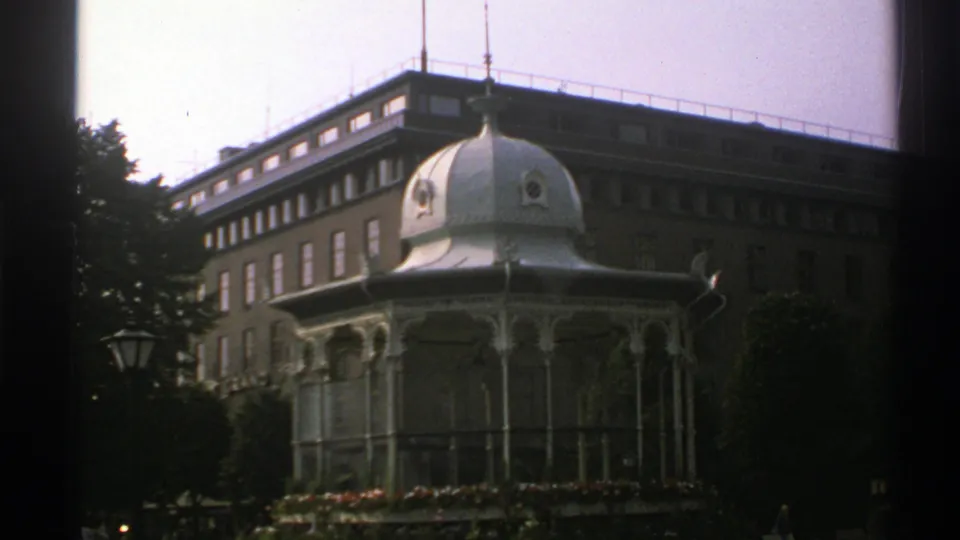 Final 1976 Norway frame holding on the white domed pavilion, with trees, a lamppost, and a large brick building in the background.