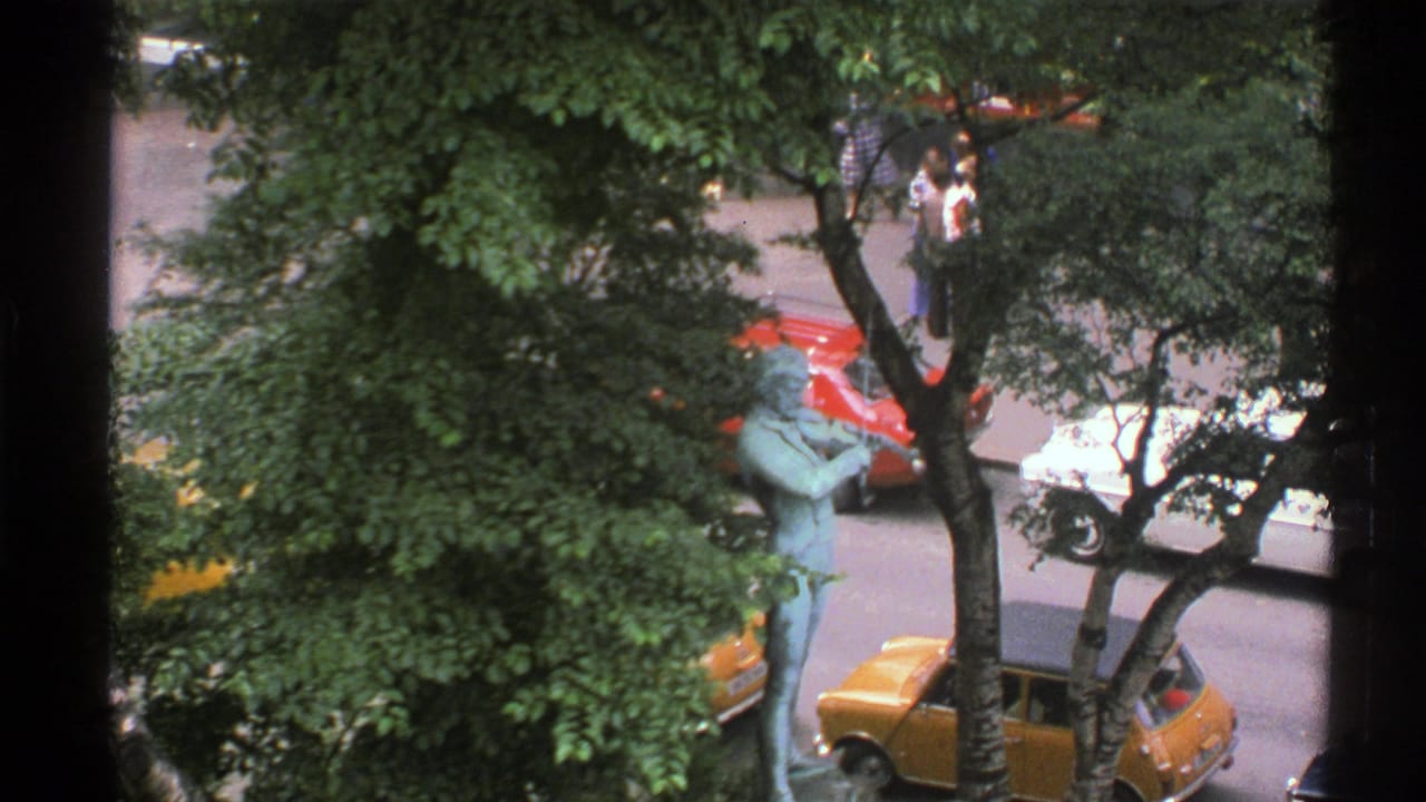 Elevated street view with trees, bronze statue, yellow car, red car, and pedestrians.