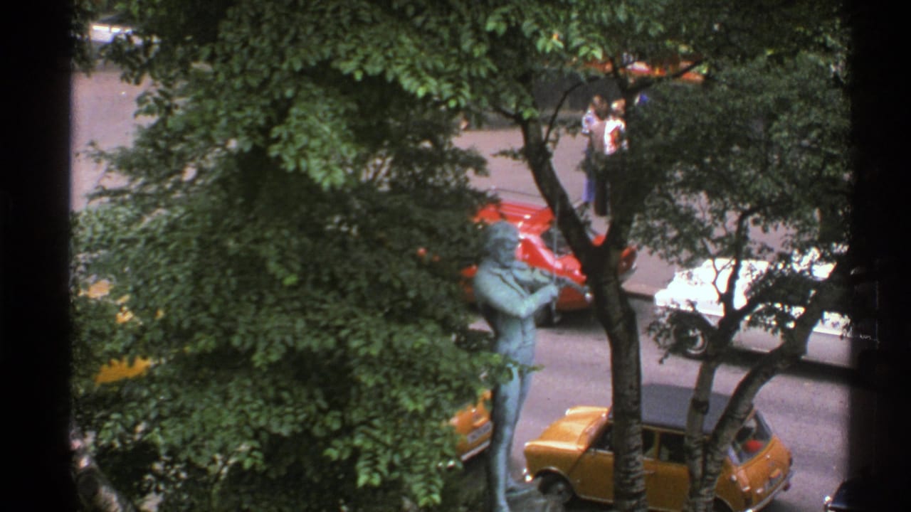 Elevated 1976 Norway street view with a bronze statue partly framed by trees, nearby cars, and pedestrians across the road.