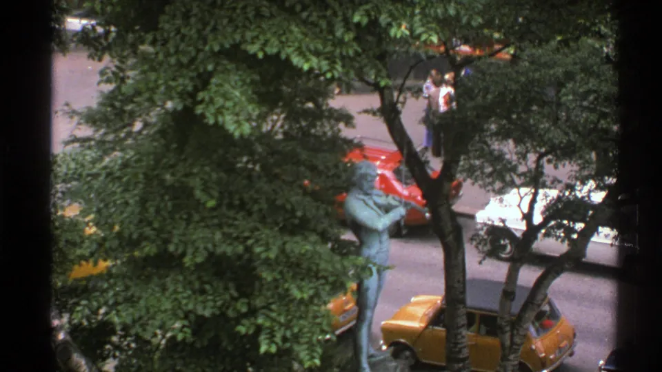 Elevated 1976 Norway street view with a bronze statue partly framed by trees, nearby cars, and pedestrians across the road.