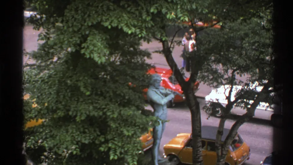 1976 Norway home-movie frame looking down through trees at a bronze musician statue, compact cars, and pedestrians on a street.